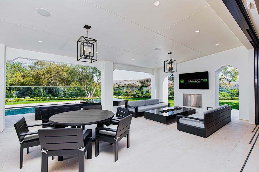  Luxurious outdoor entertainment space with table and chairs, couches, and mounted TV, lighting and speakers, and a pool in the background. 