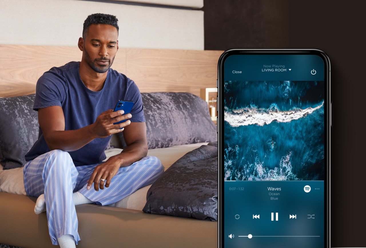  A man sitting on a bed looking at his phone with an interlay of a smartphone showing the Savant Systems music player. 