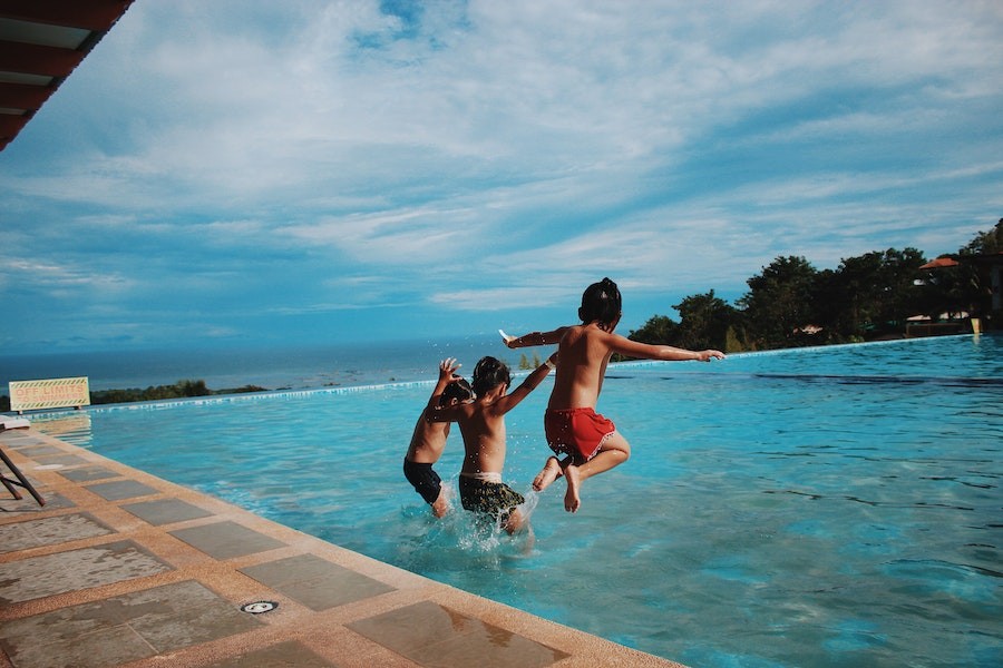 Three boys jumping into an inground pool, the ocean in the distance. 