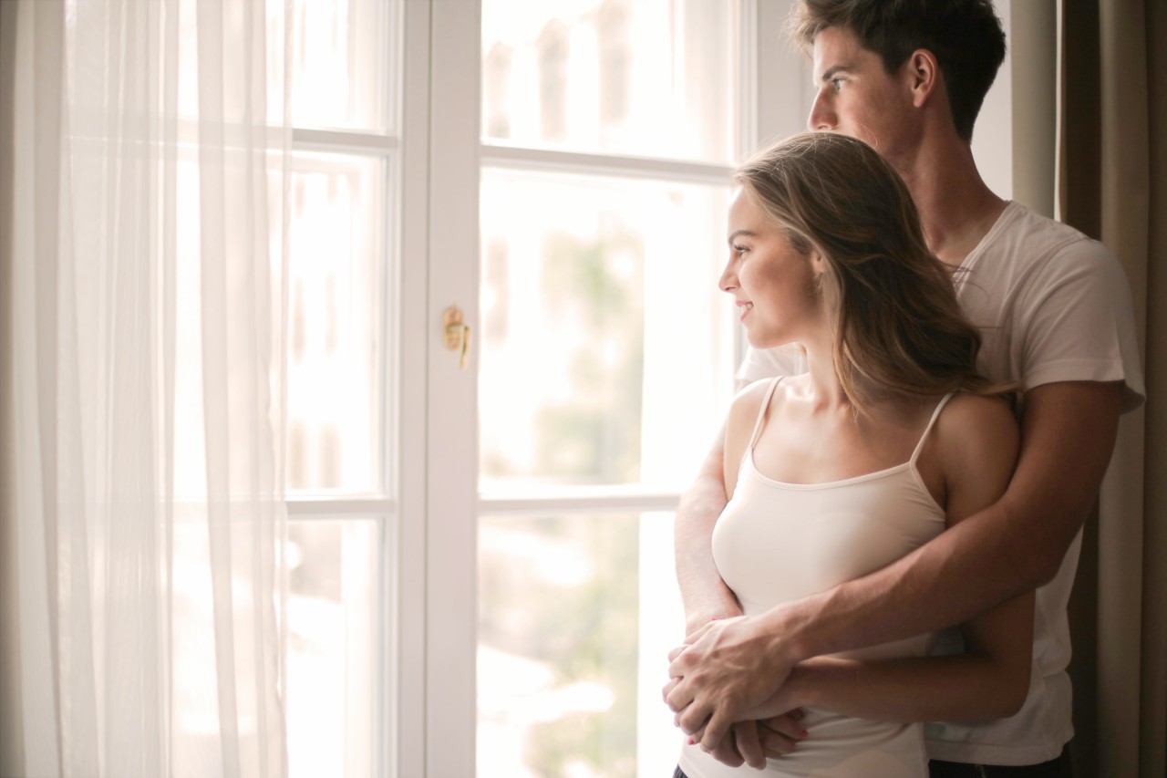 Couple standing in an embrace looking out their living room window. 