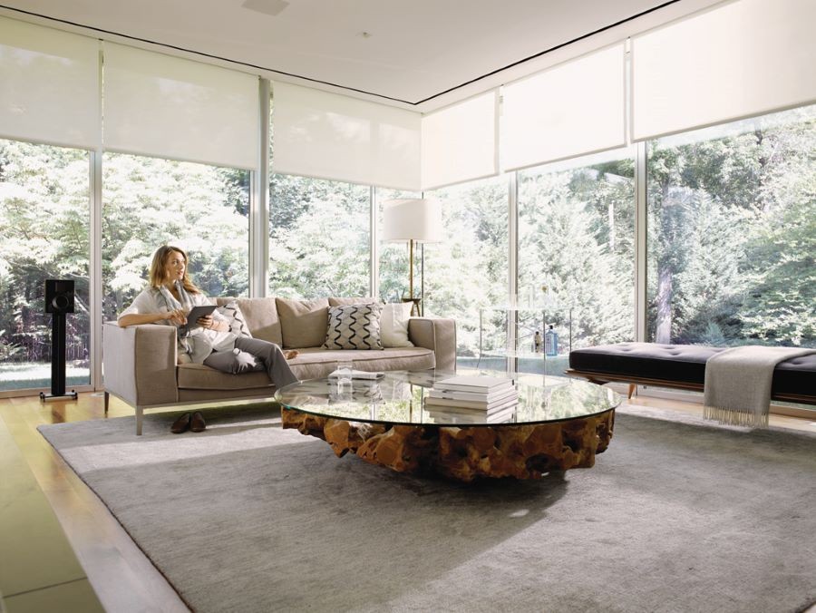 A woman sitting on a couch with a tablet in her hand, surrounded by picture windows with shades partially drawn.