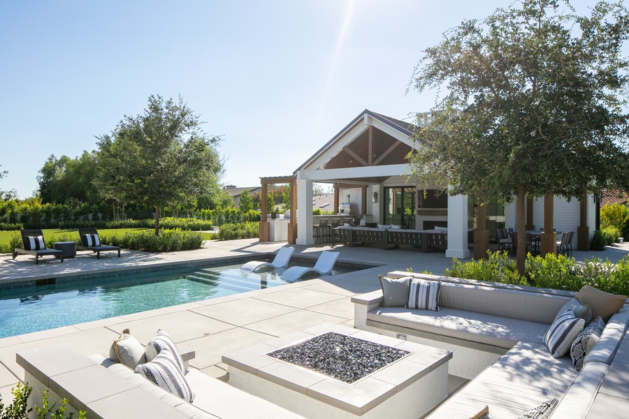 A luxury backyard with a modern pool, built-in fire pit, and stylish outdoor seating area, surrounded by lush greenery and a covered patio with an outdoor kitchen.