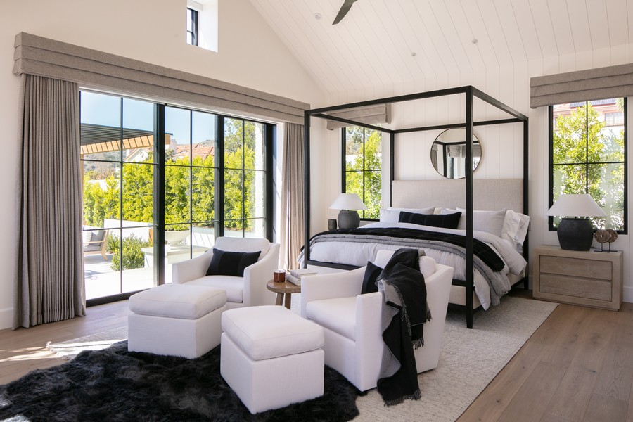 Luxury bedroom with a four-poster bed, white seating area, and floor-to-ceiling windows covered by motorized drapery and Roman shades. 